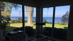 Ein Zimmer mit großem Fenster, das Blick auf eine Wiese und das Meer bietet. Ein Holzstuhl und ein kleiner Tisch mit Büchern sind vorhanden.