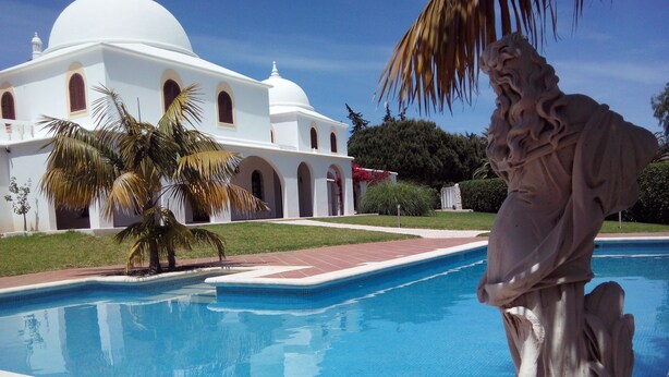 Una piscina con una estatua, palmeras y un edificio blanco con arcos al fondo.