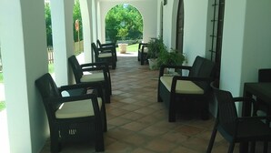 Un patio cubierto con piso de baldosas, mobiliario exterior con sillas de mimbre y cojines, y vista a un paisaje verde a través de un arco.