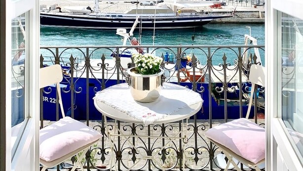 A balcony with a table and chairs, overlooking a marina with boats and buildings in the background. - Charming Sea-View Hideaway in Ortigia. Comfort, Style and Unforgettable Sunsets. (Siracusa)