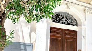 A double wooden door with a metal gate, a white wall, and a tree with green leaves. - Charming Sea-View Hideaway in Ortigia. Comfort, Style and Unforgettable Sunsets. (Siracusa)