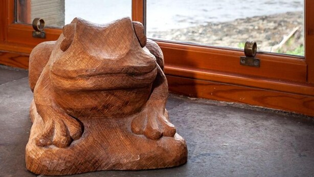A wooden sculpture of a frog on a windowsill with a view of a lake and mountains.