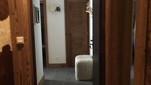 Um corredor com uma porta de madeira marcada '06', um tapete pequeno com desenho de estrela e um chão de azulejos que leva a um quarto com uma porta e um pequeno banquinho.