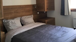Quarto com cabeceira de madeira, cama com colcha cinza, mesa de cabeceira e guarda-roupa de madeira.