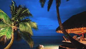 A beach scene with a thatched-roof hut, palm trees, and beach chairs.