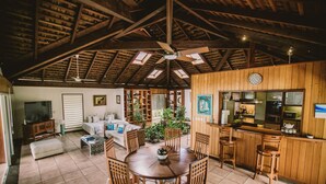 A spacious living area with a wooden ceiling, a dining table surrounded by chairs, a kitchen area with wooden cabinets, and a large window allowing natural light.