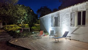 Eine Holzterrasse mit Liegestühlen und einem Tisch, umgeben von einem angelegten Garten und einem Gebäude mit Fenstern.