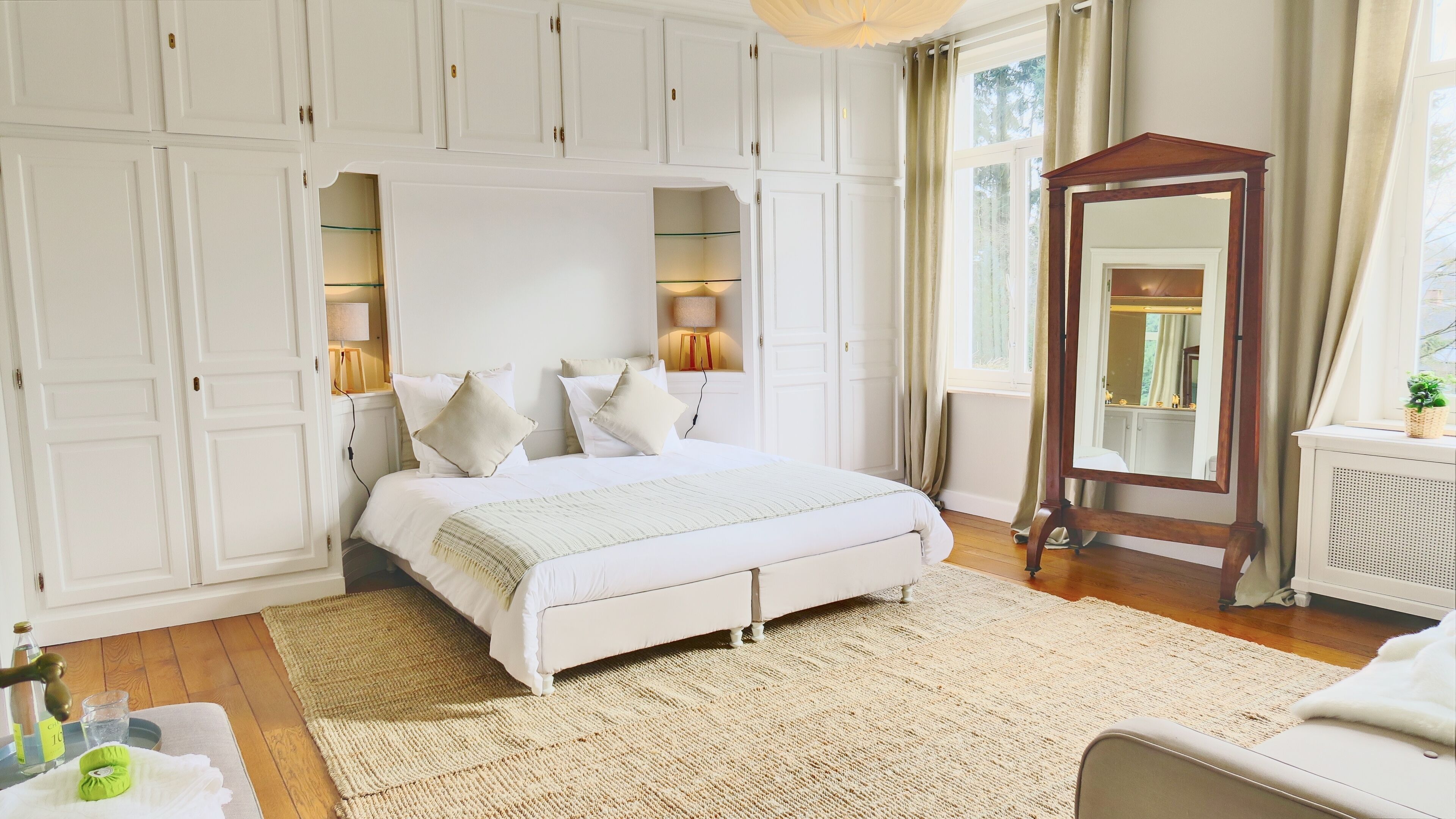 Une chambre avec un grand lit, des murs blancs et un plancher en bois. On y trouve un miroir et une petite table sur laquelle est posée une plante.