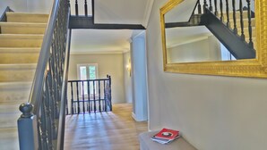 Eine Treppe mit schwarzen Spindeln und einem Holzgeländer, die zu einem Raum führt, in dem sich ein großer Spiegel und ein rotes Buch auf einem Tisch befinden.