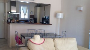 Ein Wohnzimmer mit beiger Couch, einem Esstisch mit Stühlen, einer Küchenzeile mit Kühlschrank und einem Fenster mit Vorhängen.