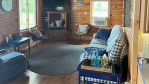 Ein gemütliches Blockhaus-Interieur mit Holzboden, einer Couch, einem kleinen Tisch mit Büchern, einem Fernseher und einem Kamin.