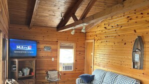 Ein gemütliches Blockhäuschen-Interieur mit einem karierten Sofa, einem Fernseher, der MLB.tv anzeigt, einer Holzbalkendecke und einem Fenster mit Jalousien.