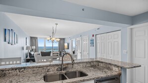 A modern kitchen with granite countertops, a stainless steel sink, and white cabinetry. There is a view of the ocean through large windows, and a chandelier hangs above the dining area.