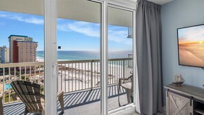 A balcony with a view of the beach and ocean, a chair, and a small table with a ship model.