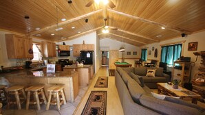 A spacious living area with a sofa, a dining table, and wooden stools. There is a kitchen with wooden cabinets and a refrigerator. - Accessible Cabin, Hot Tub, Private Dog Park & EV Charger (Big Bear Lake)