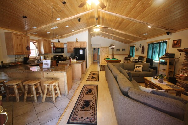 A spacious living area with a sofa, a dining table, and wooden stools. There is a kitchen with wooden cabinets and a refrigerator.