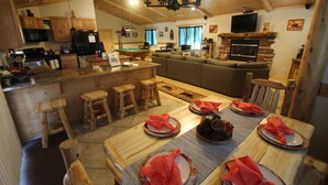 A cozy cabin interior with a dining area set for four, a living room with a fireplace, and a kitchen with wooden cabinets and a large TV. - Accessible Cabin, Hot Tub, Private Dog Park & EV Charger (Big Bear Lake)