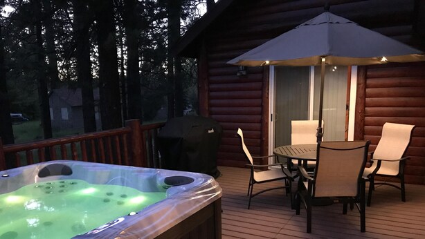 A wooden deck with a hot tub, outdoor table and chairs, and a covered umbrella. - Accessible Cabin, Hot Tub, Private Dog Park & EV Charger (Big Bear Lake)