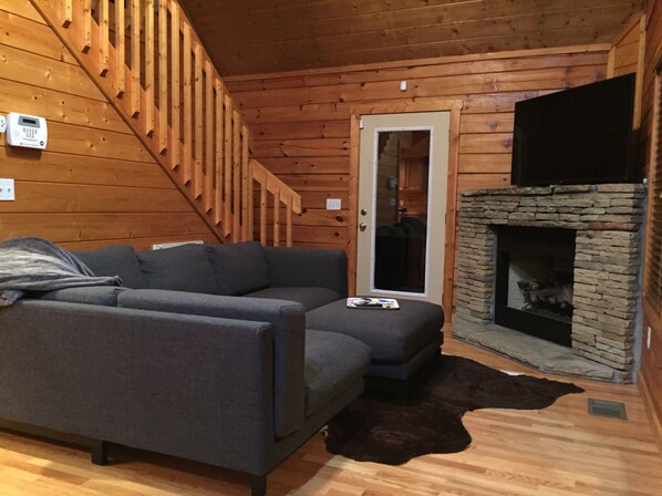 A cozy cabin interior with a gray sofa, a stone fireplace, and a wooden staircase. - Real Log Cabin w/ Secluded Hot Tub, Cozy Loft, WiFi & Close to All Attractions! (Pigeon Forge)