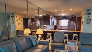 Una sala de estar moderna con un sofá, sillones y una mesa de centro. Se ve una cocina al fondo con taburetes de bar.