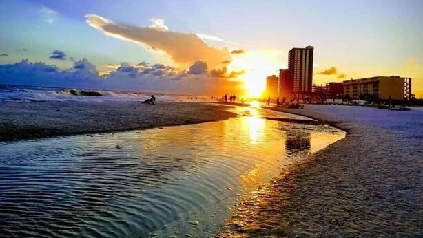 Escena de playa con un atardecer, olas y edificios al fondo.