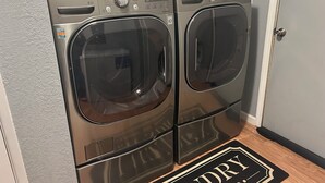 A laundry room with two front-loading LG washers and dryers, a shelf above with cleaning supplies, and a black mat on the floor with "Laundry" written on it. - Black Gold Getaway .....Walking distance to town (Valdez)