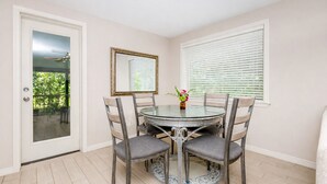 Un comedor con una mesa redonda de vidrio, cuatro sillas grises tapizadas, un jarrón con flores, un espejo en la pared y una ventana con persianas.