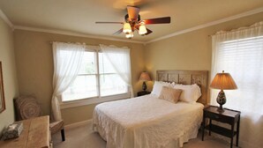A bedroom with a large bed, two bedside lamps, a ceiling fan, and a window with sheer curtains.