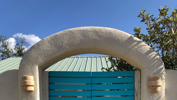 A turquoise gate with a metal handle, set within a white arched structure, leading to a building with a green roof.