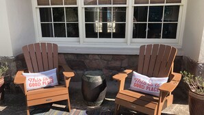 Two wooden Adirondack chairs with pillows that read "This is a good place" placed outside a building with a stone wall and a window with blinds.