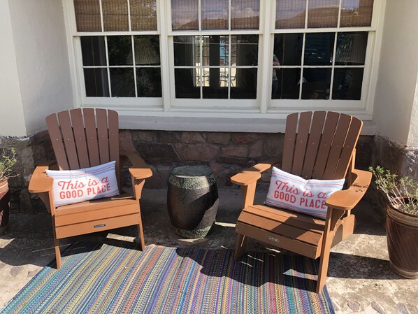 Two wooden Adirondack chairs with pillows that read "This is a good place" placed outside a building with a stone wall and a window with blinds.