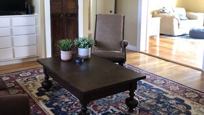 A living room with a dark wooden coffee table, a patterned rug, a wicker chair, and a television on a white cabinet.