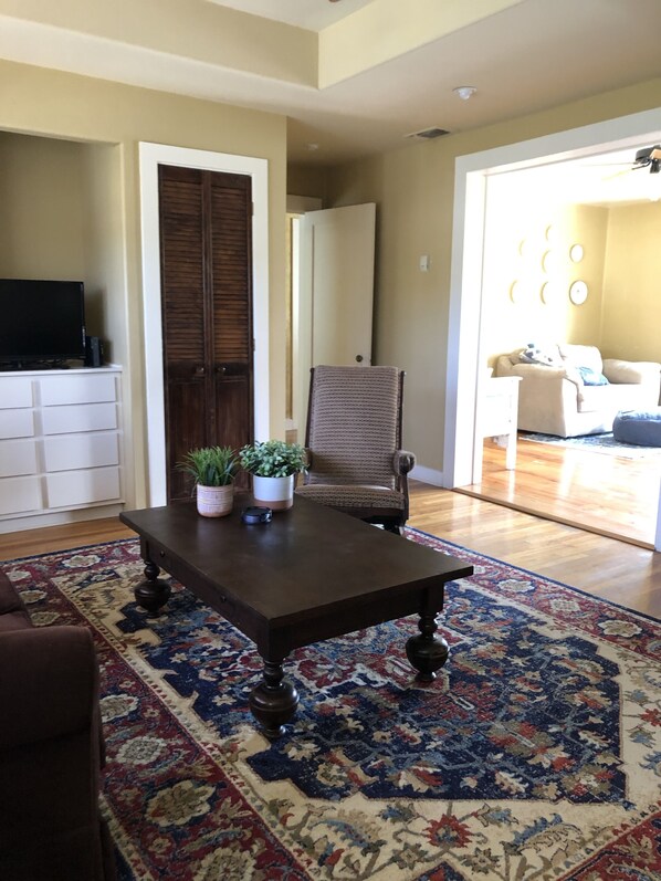 Una sala de estar con una mesa de centro de madera oscura, un tapete estampado, un sillón de mimbre y un televisor sobre un mueble blanco.