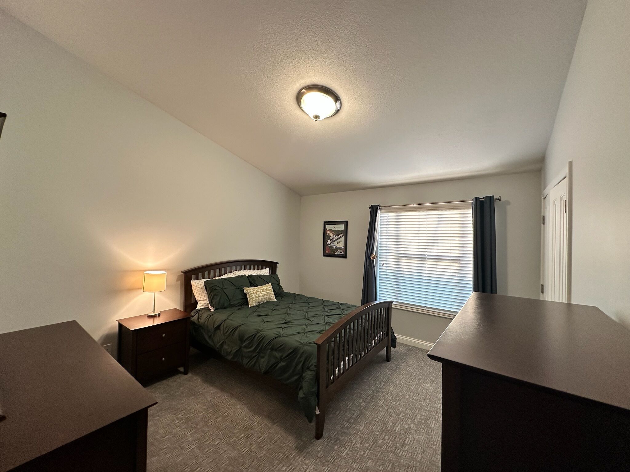 A bedroom with a single bed, a nightstand, a desk, a window with blinds, and a ceiling light.
