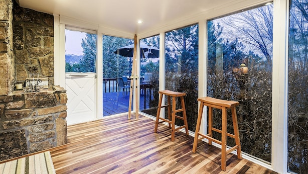 Una habitación con chimenea de piedra, piso de madera y grandes ventanales con vistas a una terraza y árboles.