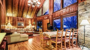 Un interior de madera acogedor, con zona de comedor, sofá y ventanas amplias que dan a unas arboledas.