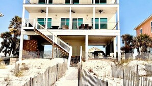 A multi-story beachfront house with a covered front porch, a staircase leading to the entrance, and a sandy beach area with a wooden fence. - Gulf front home on North Cape, outdoor kitchen, elevator @ lots of beach gear (Port St Joe)