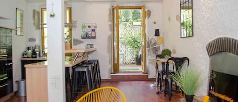 A spacious living area with a wooden floor, a yellow wicker chair, a fireplace, and a kitchen area visible through an open doorway.