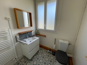 Une petite salle de bain propre, avec un lavabo, un miroir et des toilettes blancs. Il y a une fenêtre au-dessus des toilettes et un radiateur sur le côté gauche.