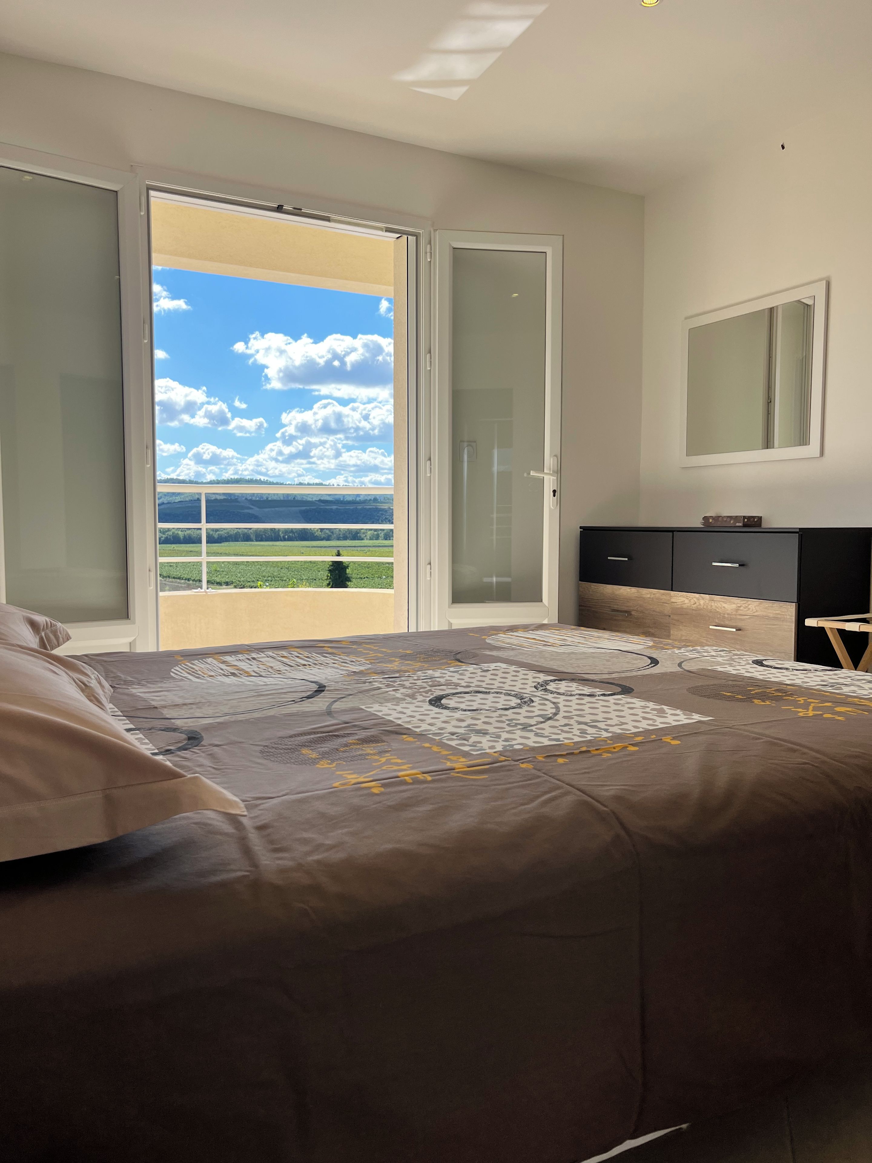 A bedroom with a bed, a dresser, a mirror, and a sliding glass door leading to a balcony with a view of greenery and a blue sky.