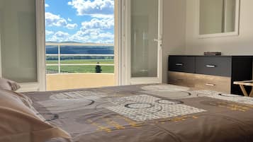 A bedroom with a bed, a dresser, a mirror, and a sliding glass door leading to a balcony with a view of greenery and a blue sky.