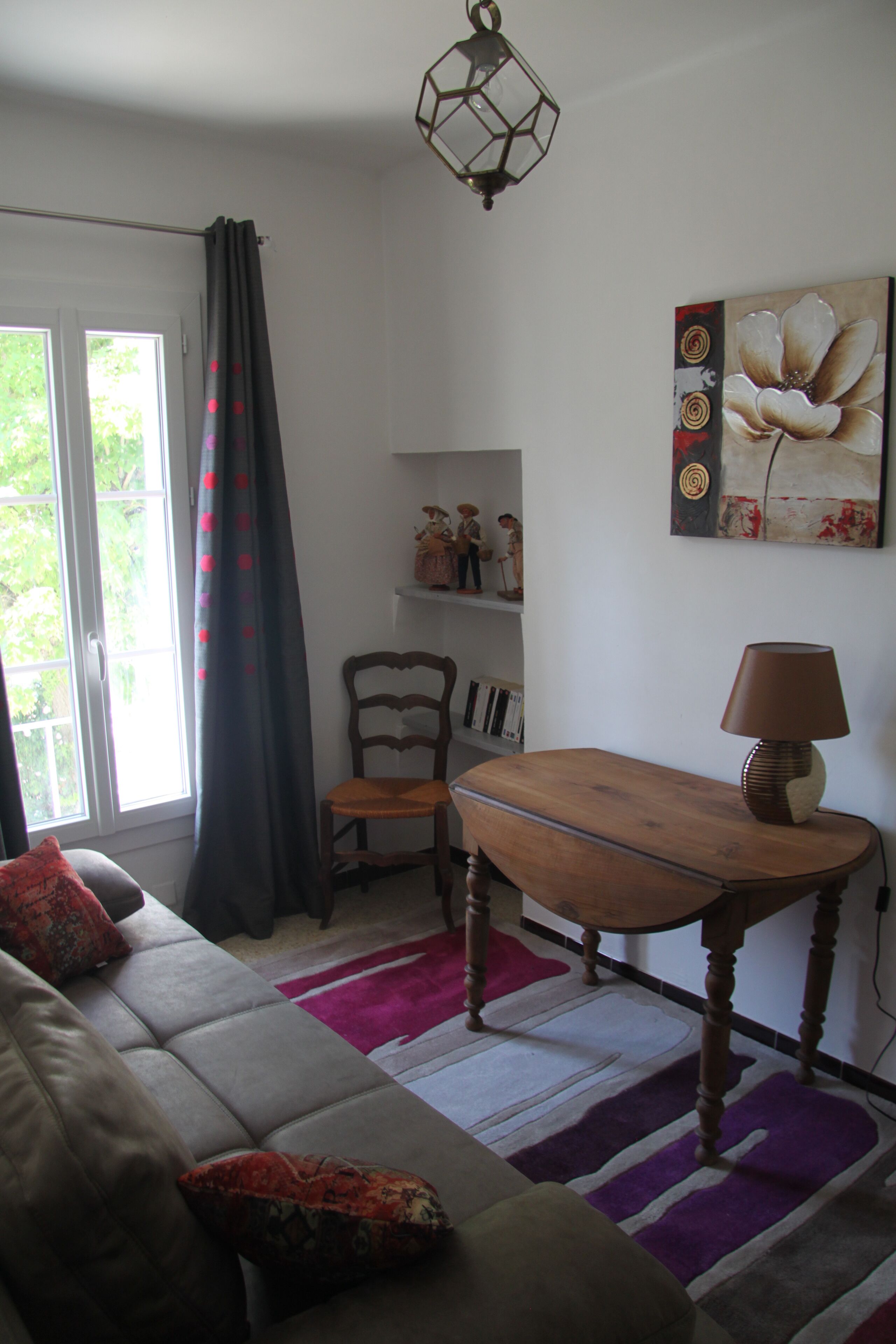 A living room with a sofa, a wooden table, a chair, a lamp, a bookshelf, and a window with curtains.