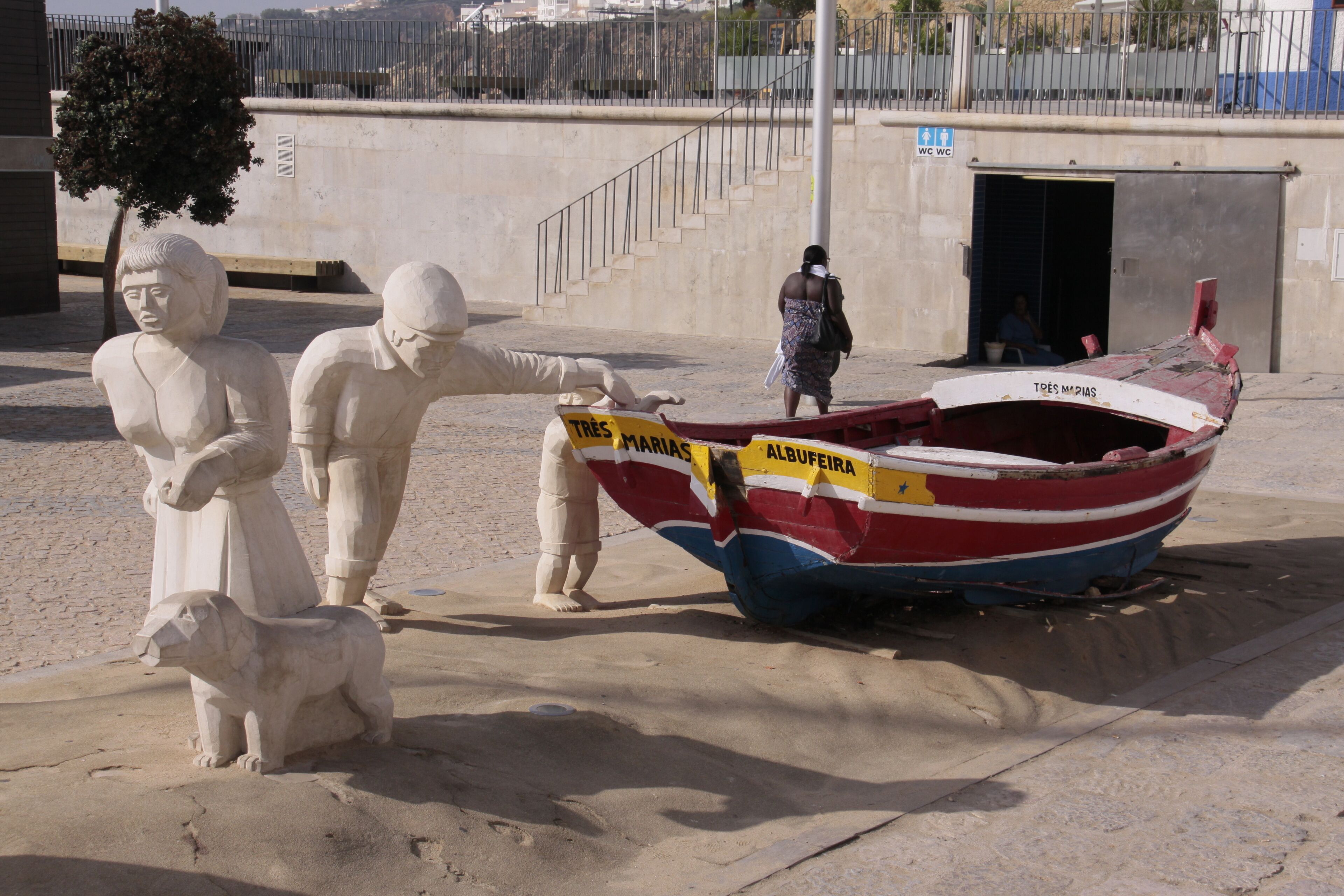 A sculpture of a man and a woman with a dog, a boat with 'TRÉS MARIAS ALBUFÉIRA' written on it, and a staircase leading to a building.