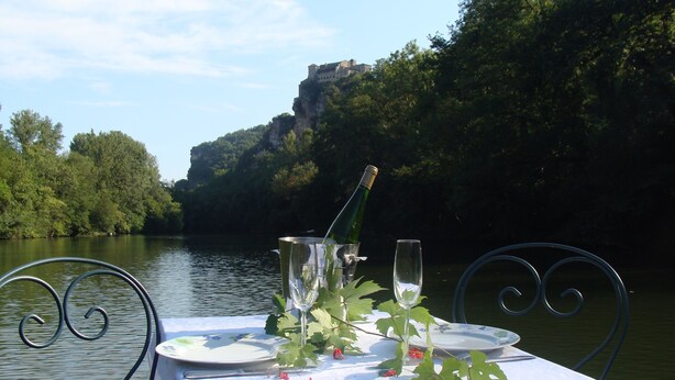 Een gedekte tafel voor twee met champagne, glazen en borden op een witte tafelkleed, langs een rivier met een kasteel op de achtergrond.