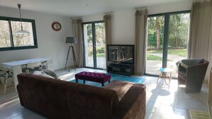 Een woonkamer met een bruine bank, een paars voetenbankje, een televisie en een eetgedeelte met een witte tafel en stoelen.