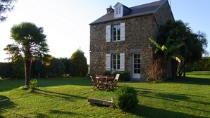 Une maison en pierre avec une pelouse bien entretenue, une terrasse avec une table et des chaises, et un mât pour drapeau.