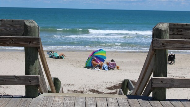 A wooden beach deck leading to a sandy beach with a colorful umbrella and beach chairs. - 4 BEDROOM HOUSE JUST A WALK TO THE BEACH! (Indian Harbour Beach)