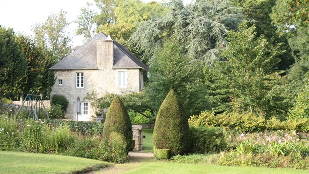 A stone house with a sloped roof, surrounded by a well-maintained garden with various plants and trimmed hedges. - Charming house in the park of a historic monument castle (Barbeville)