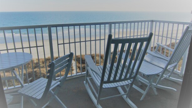 A balcony with white metal chairs and a small round table, overlooking a beach with the ocean in the background.