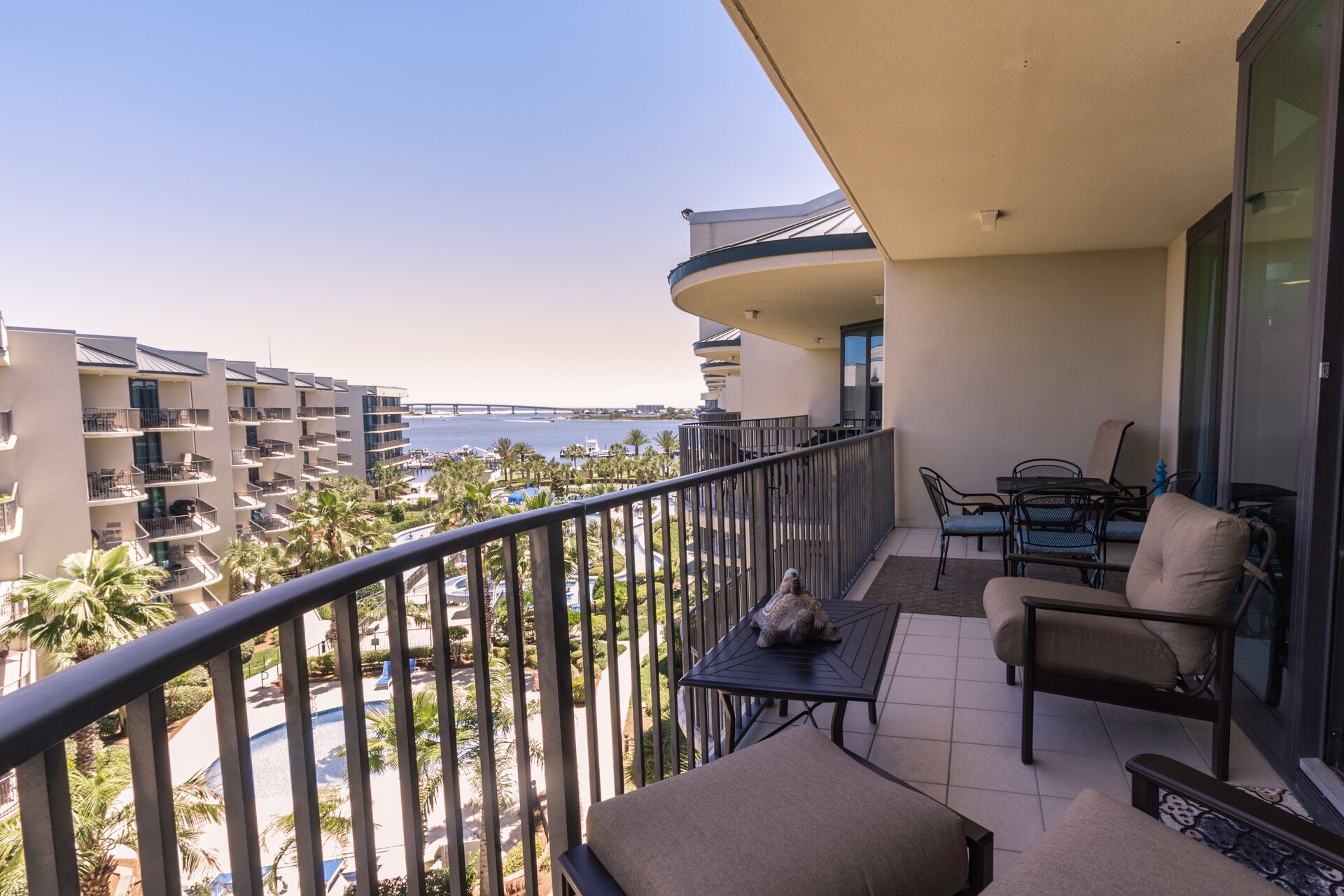 Un balcón con vistas al mar y palmeras, una zona de comedor con sillas y una mesa, y una piscina al fondo.